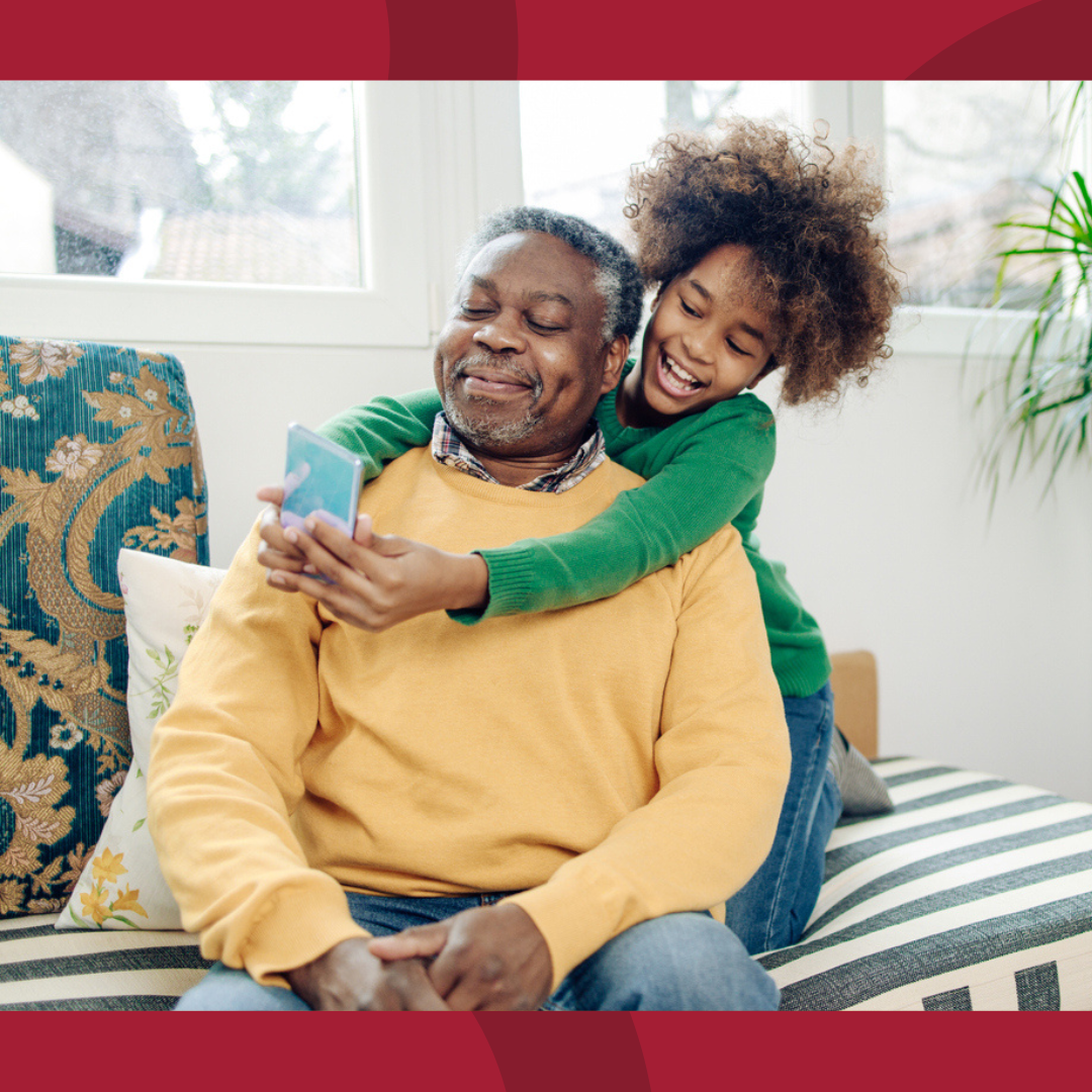 A grandfather is sitting and smiling while his young granddaughter has her arms around him from behind and showing him something on her phone.